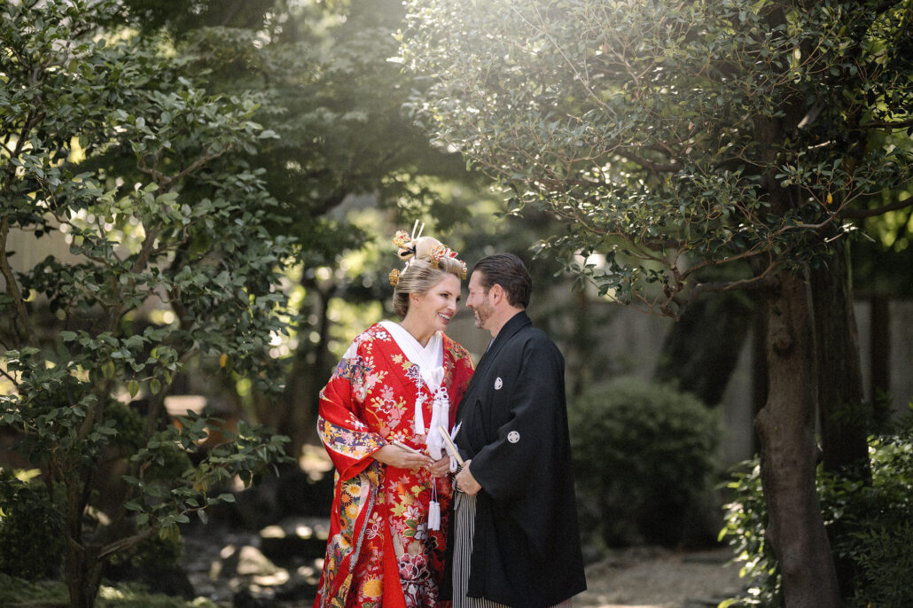 Bride in a red Japanese wedding kimono and groom in black montsuki and striped hakama smile at each other under trees in a Tokyo garden