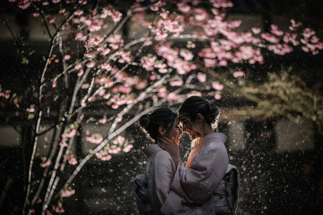 At dusk two brides in kimono touch foreheads under pink blossom as drops of water catch the backlight in a Japanese garden.