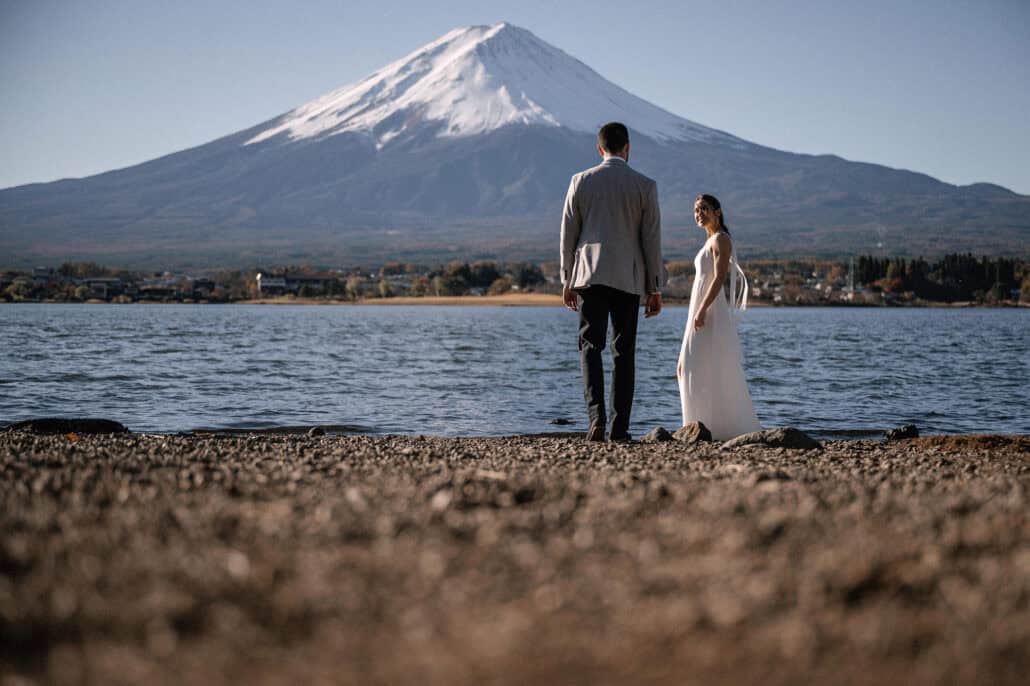 A couple stand on the pebble shore of Lake Kawaguchiko, looking up across the water at a clear, snowy Mt Fuji.