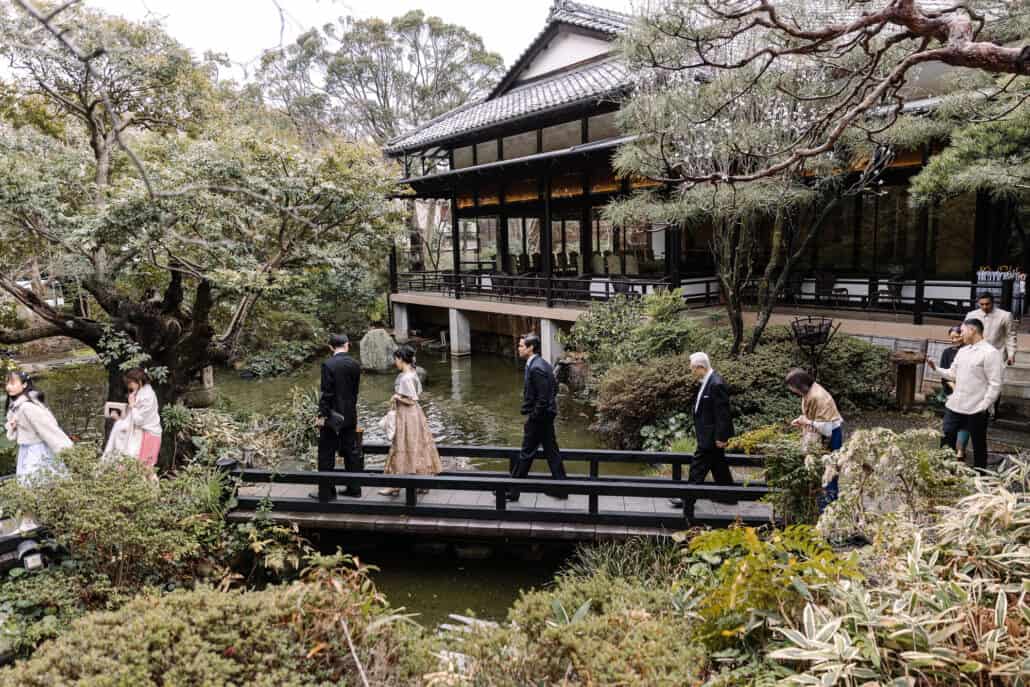 Wedding guests cross a wooden bridge over the garden pond at Motoyu Jinya, the old wooden hall and pines behind them.