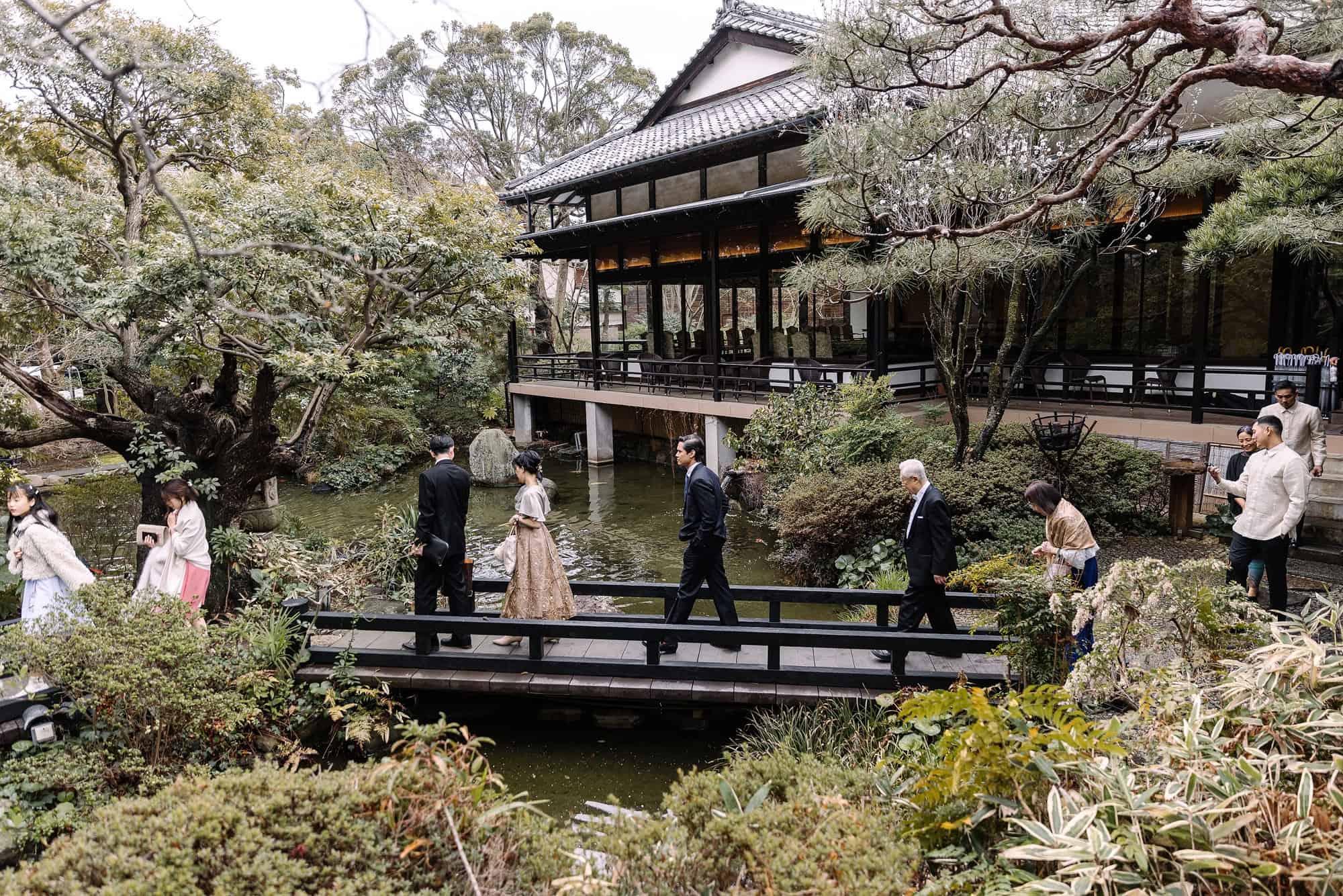 Wedding guests cross a wooden bridge over the garden pond at a ryokan in Tsurumaki Onsen, an old timber hall and pines behind them.