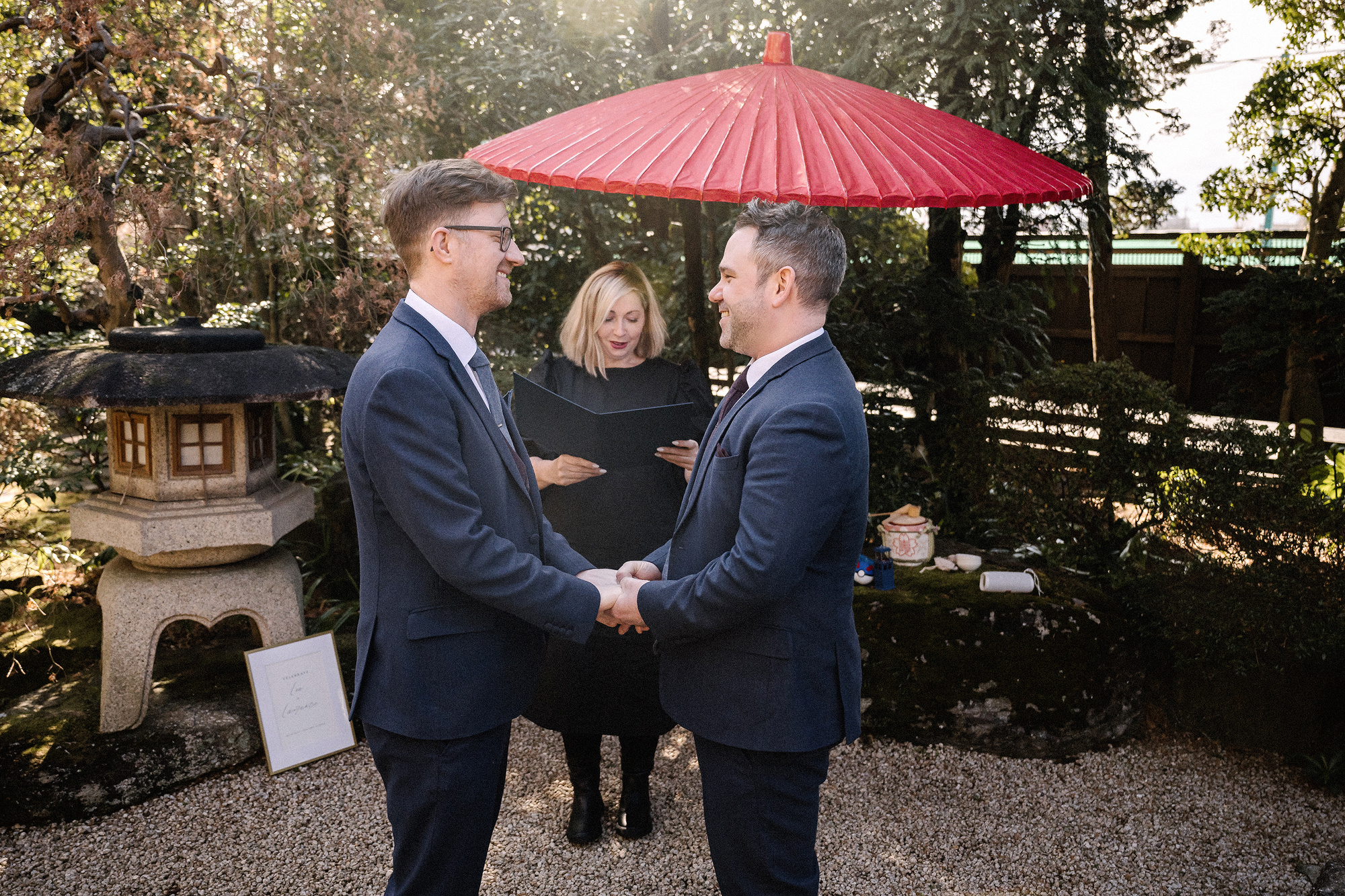 Two grooms hold hands for their ceremony under a large red parasol beside a stone lantern in a winter garden in Kawagoe, a wedding in Japan.