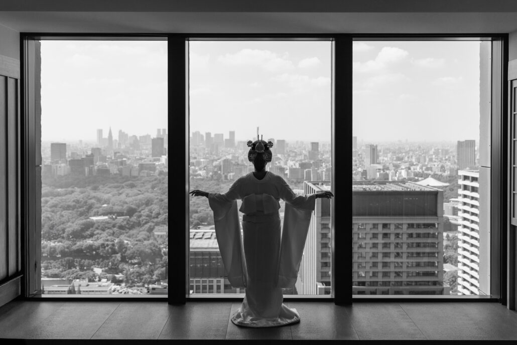 Black and white silhouette of a bride in kimono and nihongami, arms raised, before floor-to-ceiling windows high above the Tokyo skyline