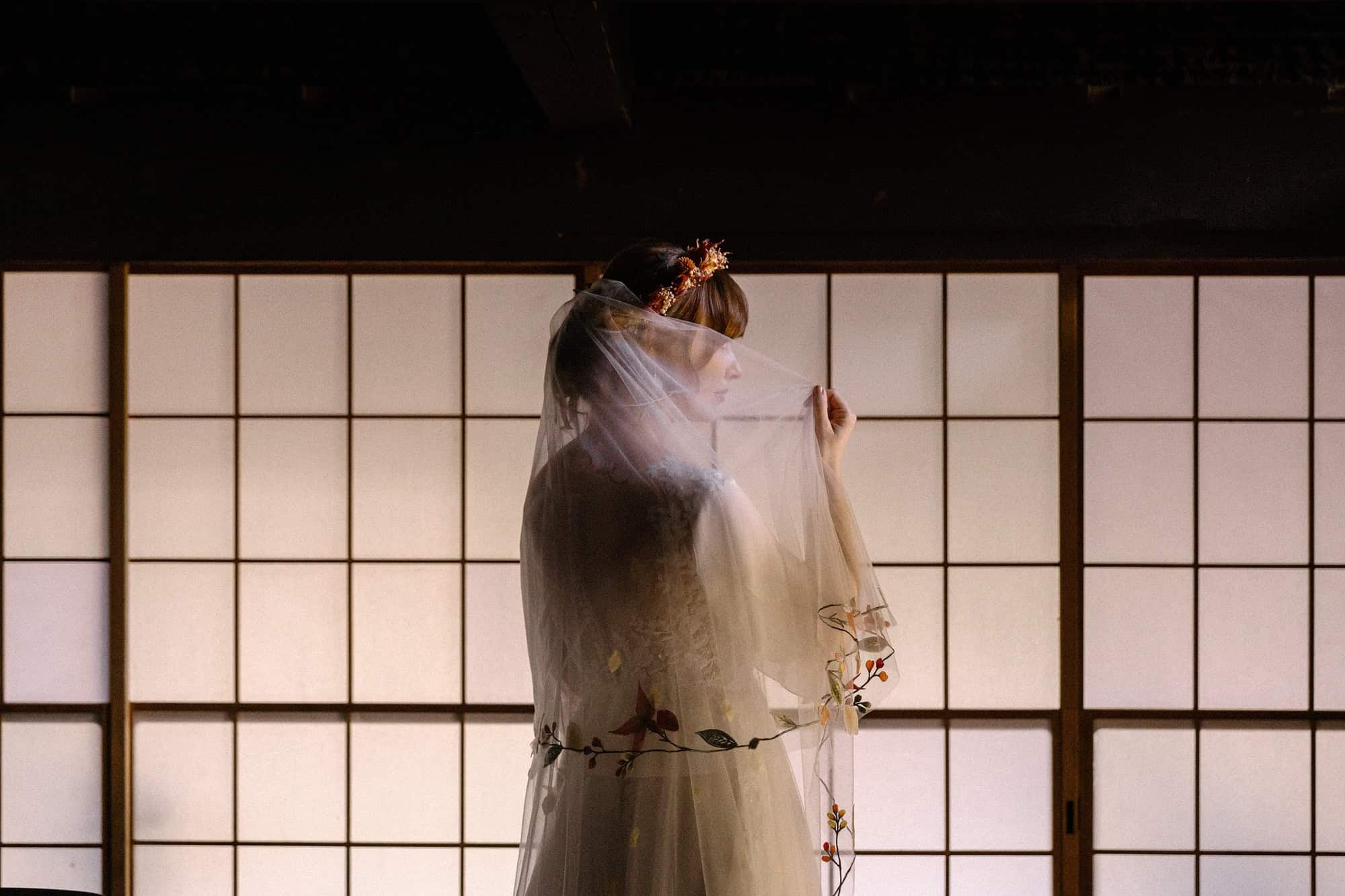 Against glowing shoji, a bride lifts the edge of her veil scattered with autumn leaves, in an old mountain house for her wedding in Japan.