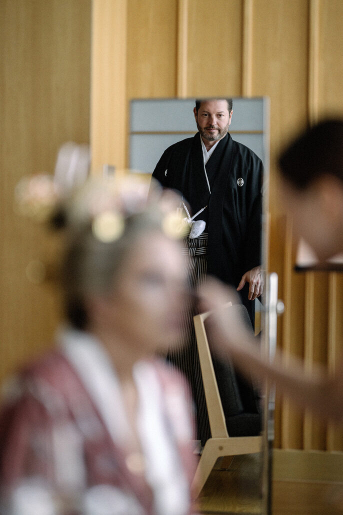 Groom in black montsuki and striped hakama, seen in a mirror, waits while his bride has her makeup done before their Tokyo wedding