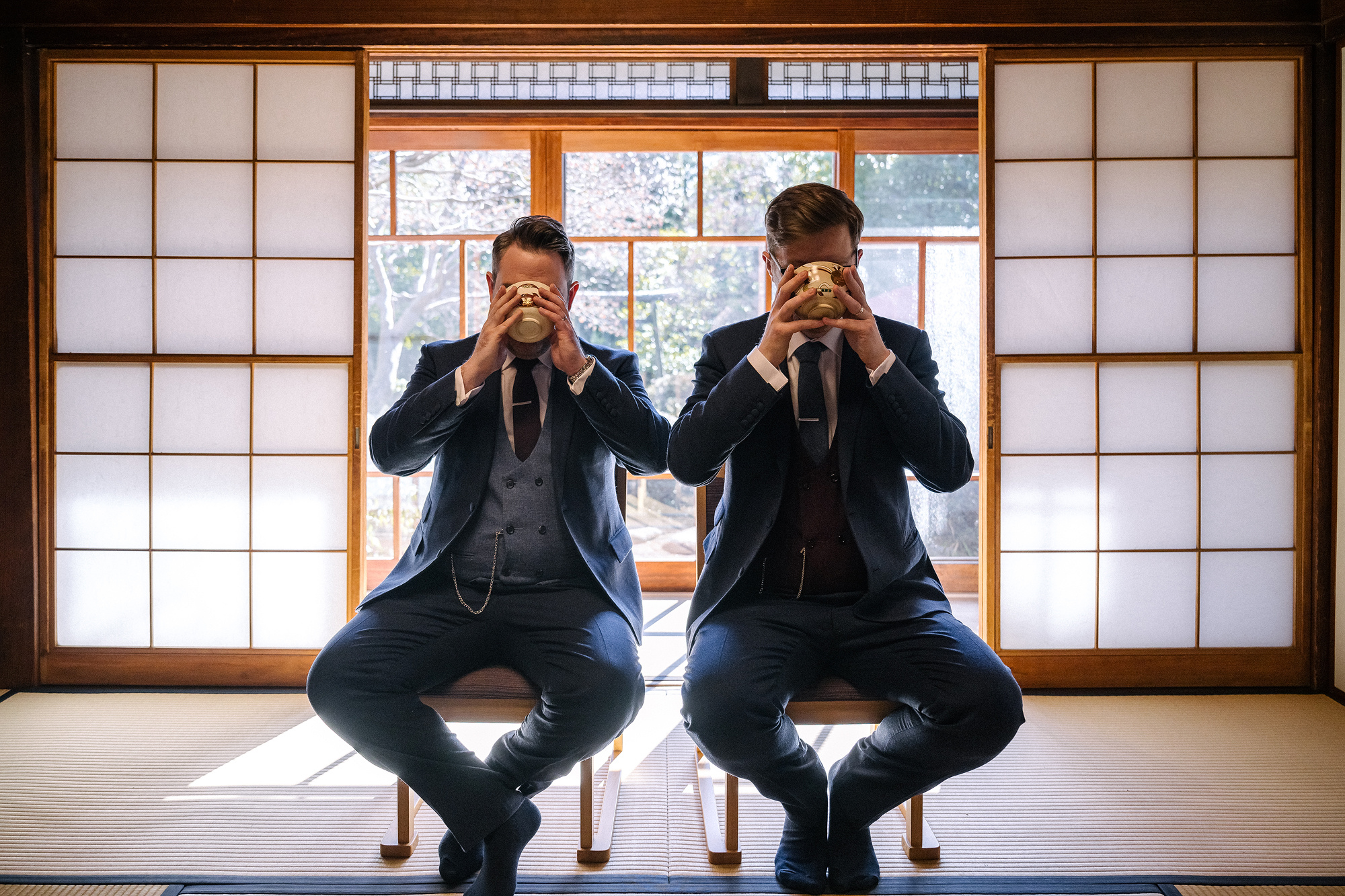 Two grooms drink matcha in the same moment against glowing shoji at Tantoku in Kawagoe, a tea ceremony wedding in Japan.