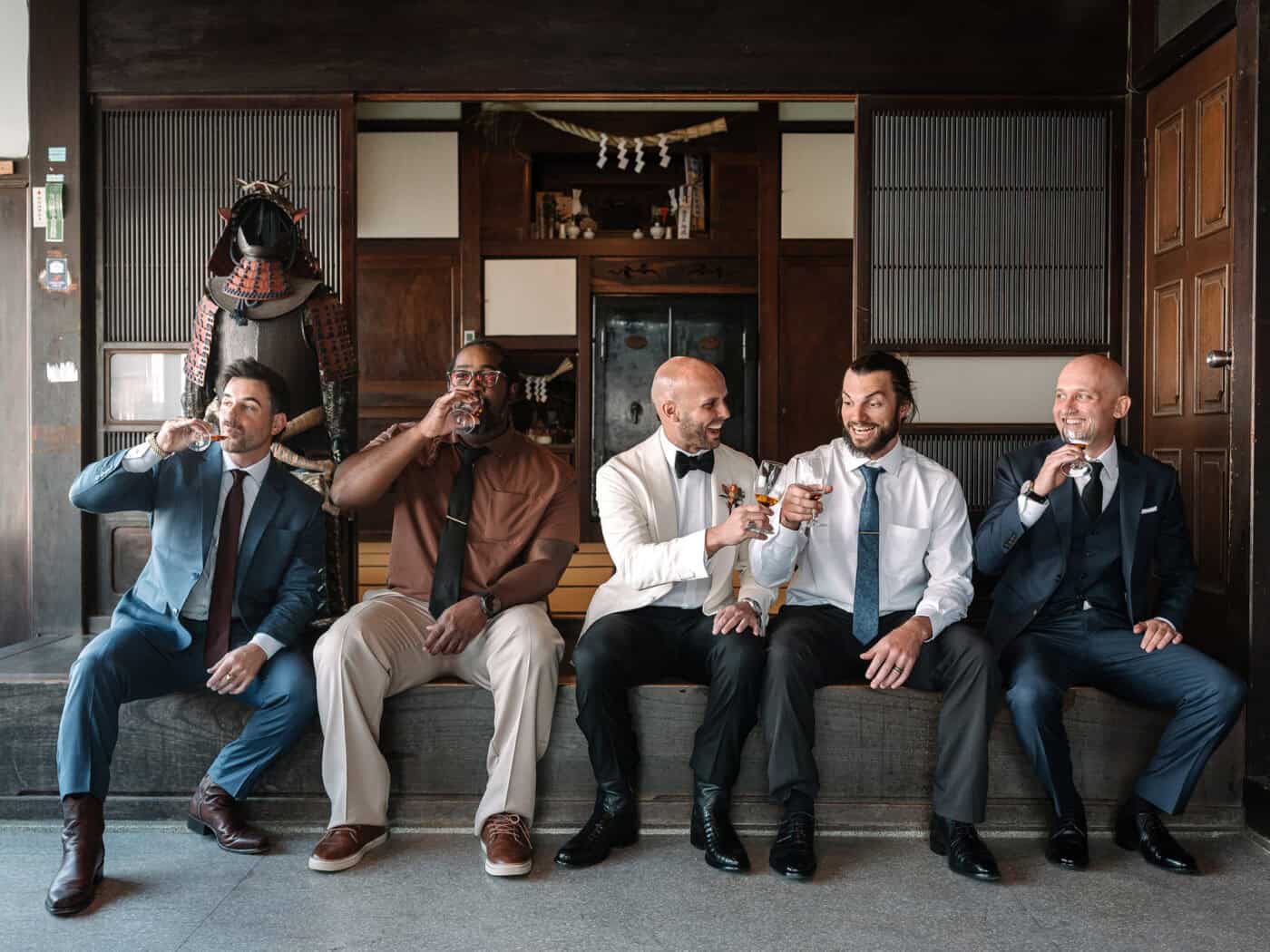 The groom and his friends sip whisky on a long bench beside a suit of samurai armour in the wood panelled front room of the house.