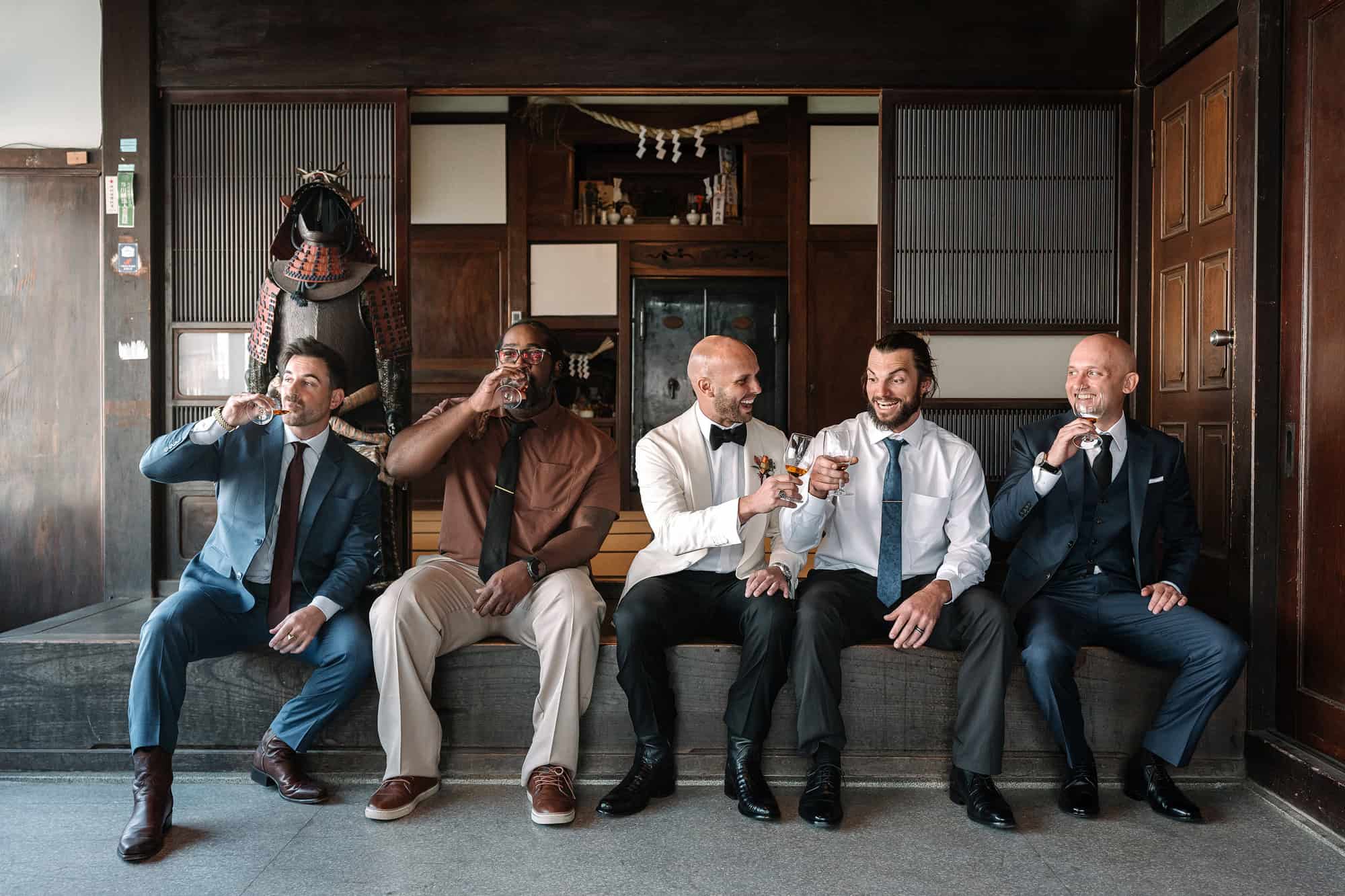 The groom and his friends sip whisky on a long bench beside a suit of samurai armour in the wood panelled front room of the house.