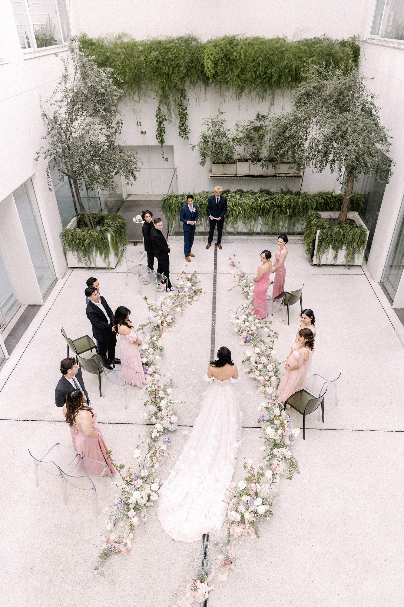 From above, a bride walks a winding aisle of low pink and white flowers through a white courtyard, a garden wedding ceremony in Tokyo.