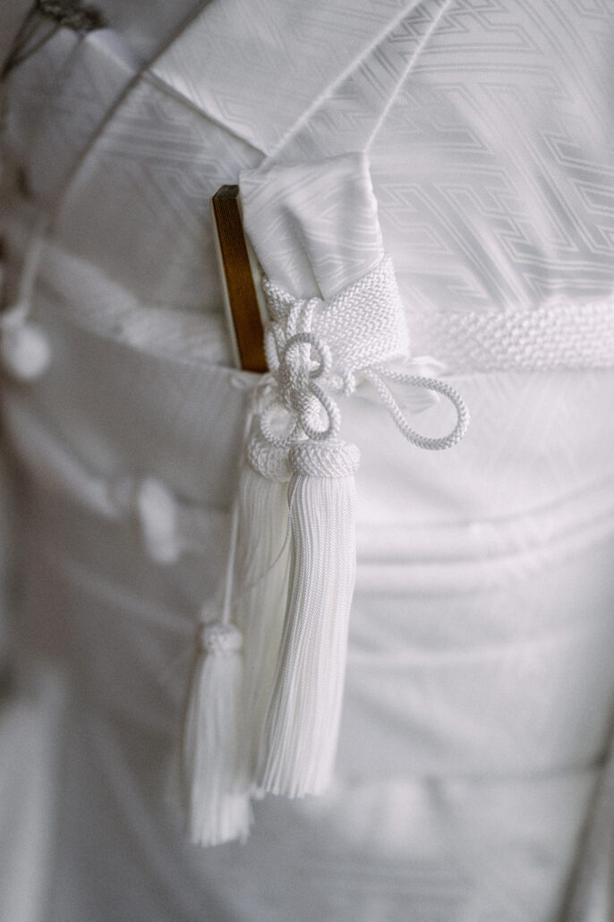 Close-up of white silk tassels and a knotted braided cord tucked into the folds of a white Japanese wedding kimono on the bride