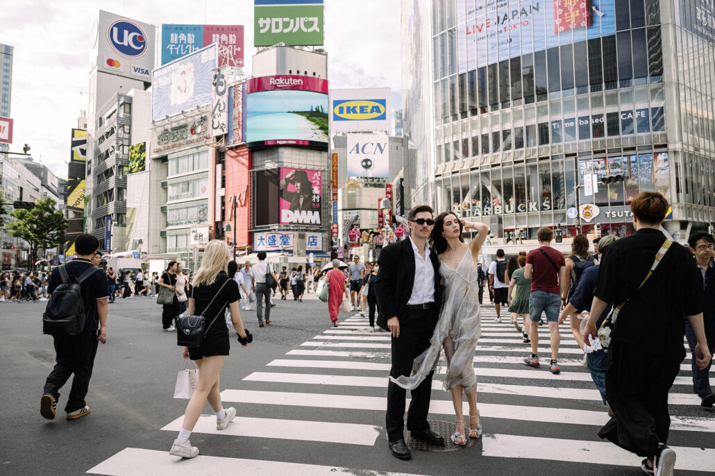 A wide view of Shibuya Crossing in Tokyo, a couple small in the middle of the crowd beneath the billboards.