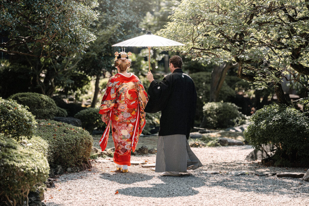 Groom holds a white parasol over his bride in a red iro-uchikake and nihongami as they walk a gravel path through a Tokyo garden