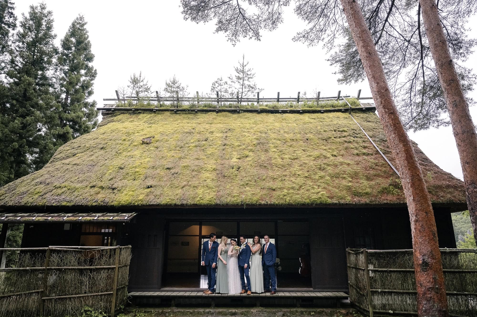 A wedding party of six stands in the doorway of a moss-covered thatched house between tall pines, an intimate wedding in Japan near Lake Saiko.
