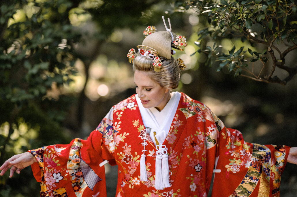 Bride in nihongami with red and gold flower kanzashi, wearing a red iro-uchikake with white tassels, sleeves spread among garden trees