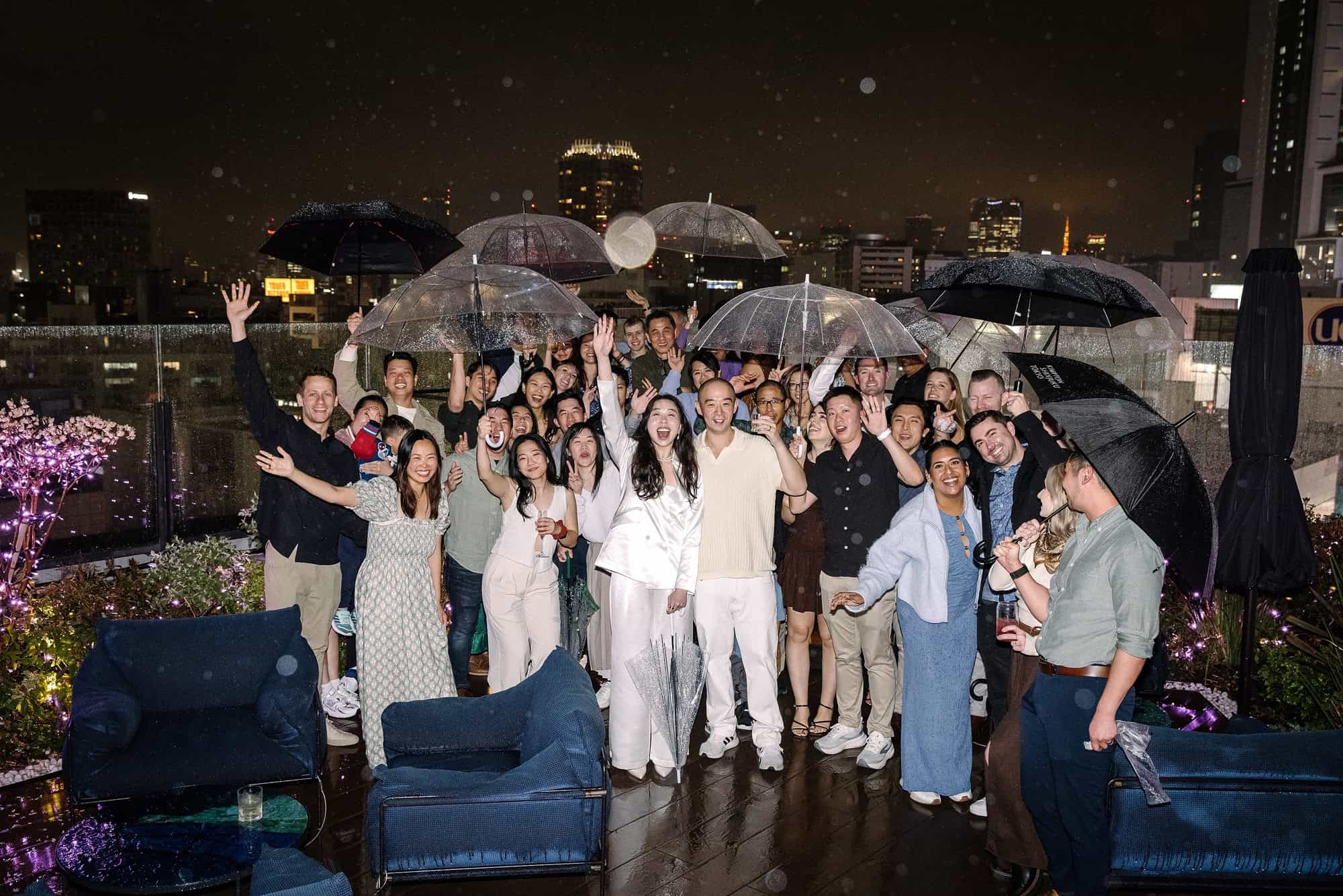 Wedding guests crowd together under clear umbrellas on a rainy Shibuya rooftop at night, cheering on the first evening of a wedding weekend in Japan.