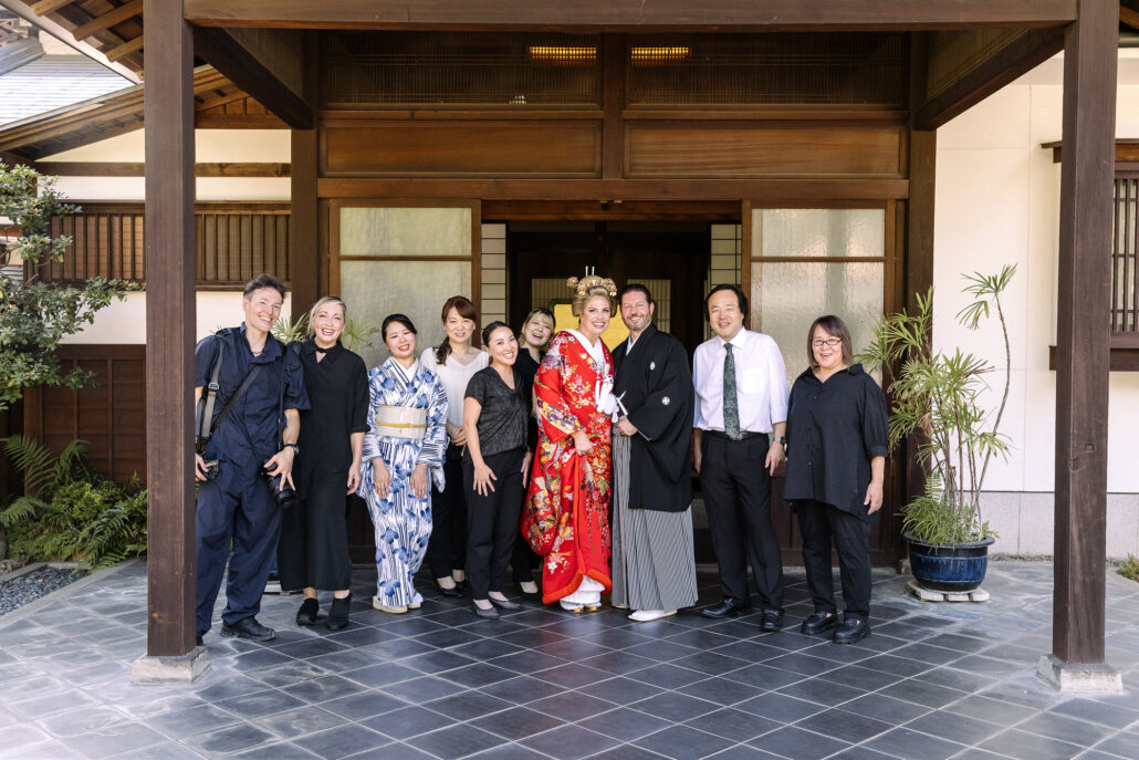 Bride in a red iro-uchikake and groom in hakama stand with the wedding team at the wooden entrance of a traditional Japanese house