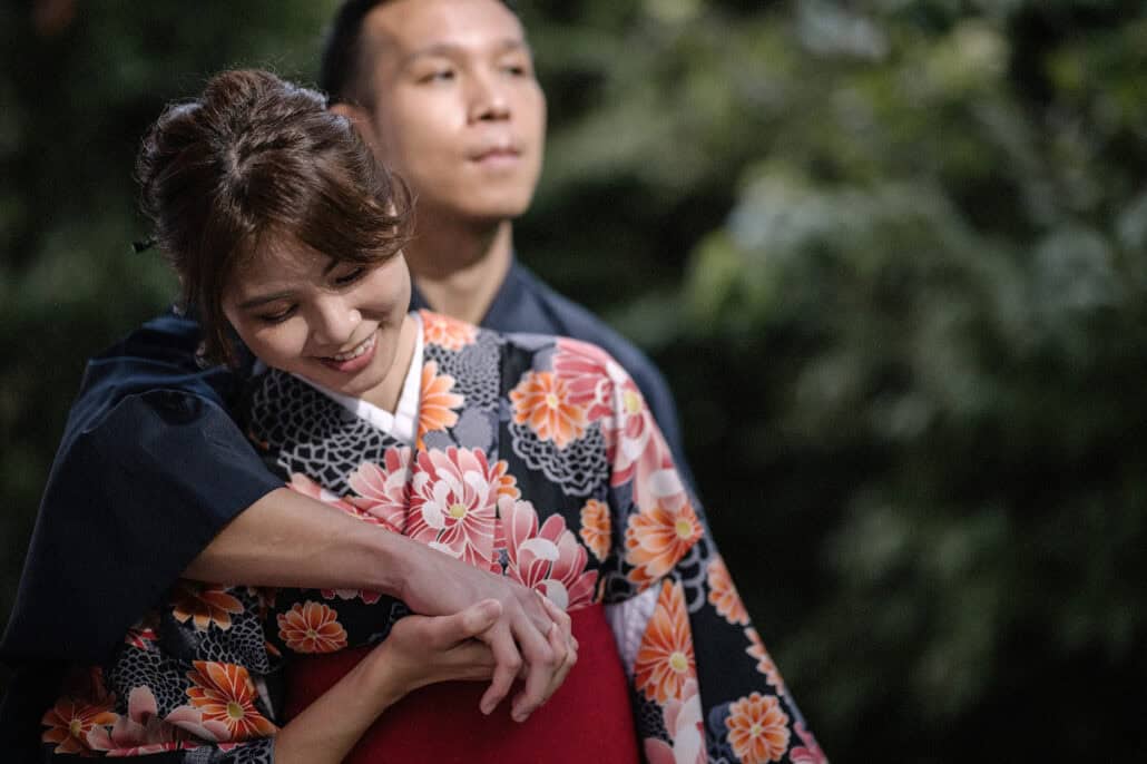 He wraps his arms around her from behind as she smiles down, her kimono printed with pink and orange chrysanthemums.