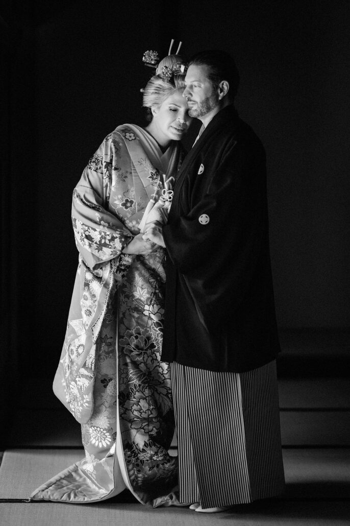 Black and white portrait of a bride in nihongami and a richly patterned uchikake leaning into her groom in crested kimono and hakama
