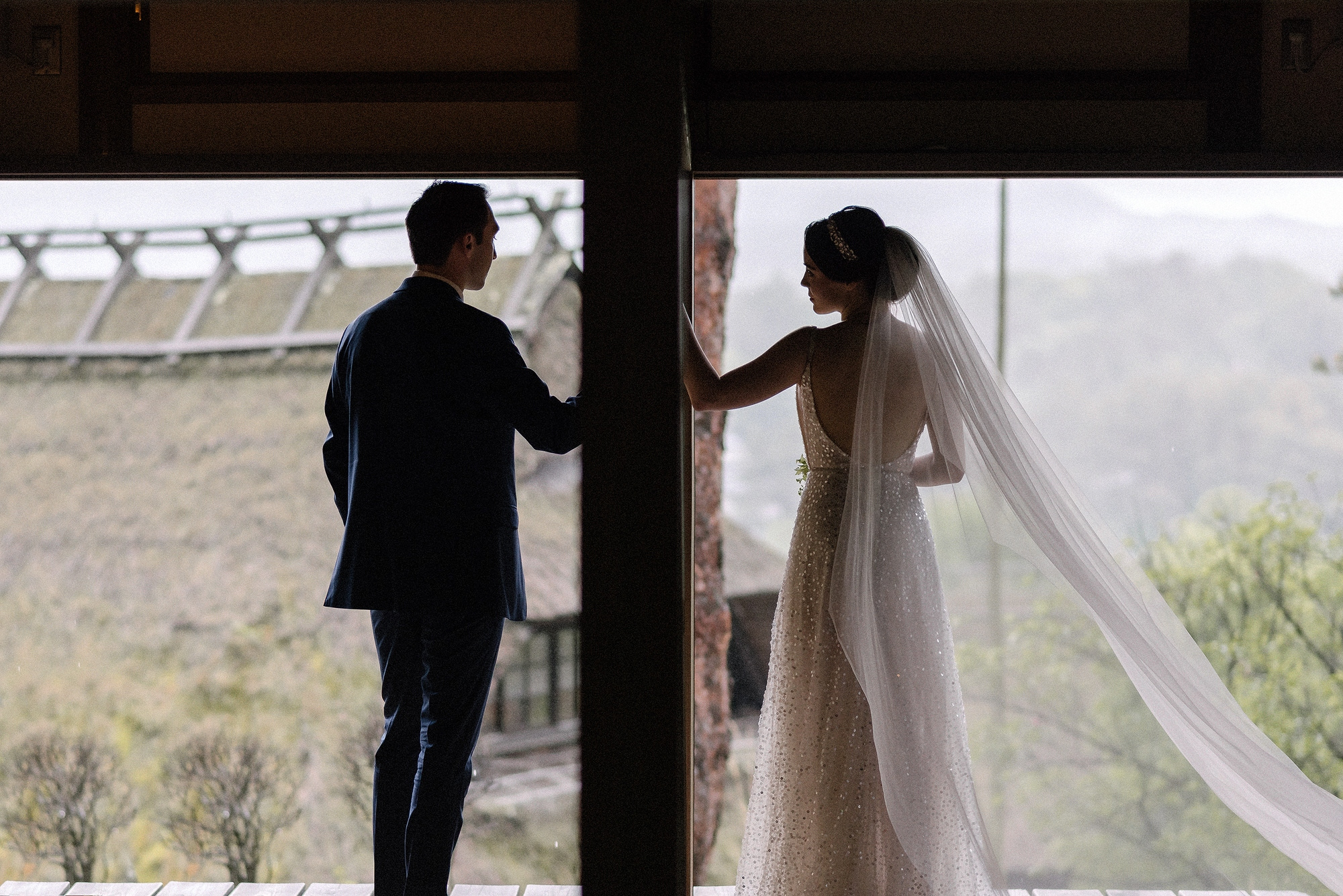 A bride and groom look out from an open doorway over thatched roofs and misty hills, her veil lifting, on a rainy wedding day near Mt Fuji in Japan.
