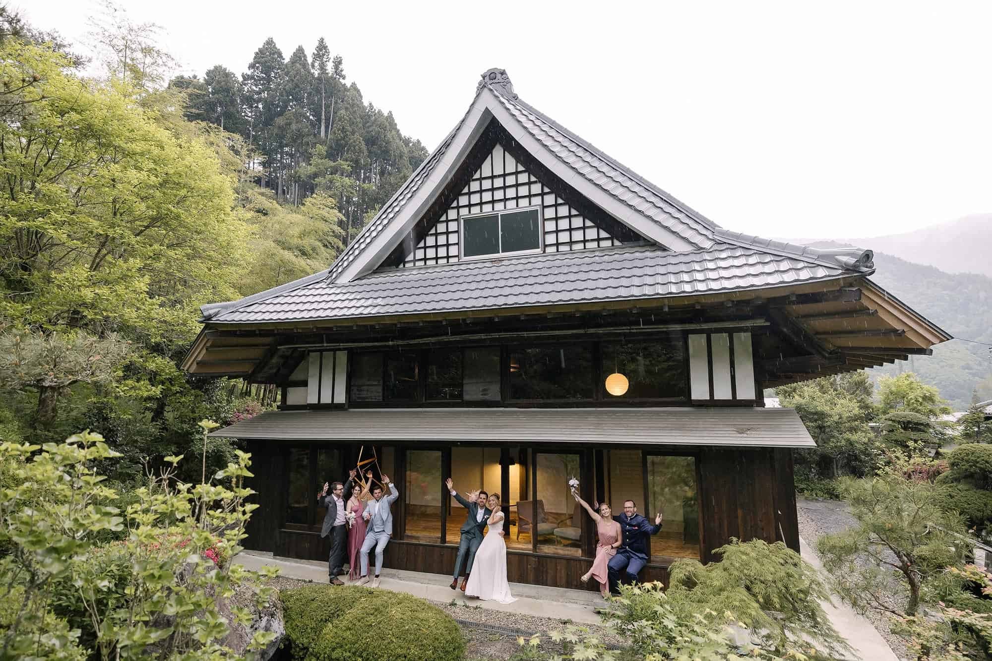 Friends cheer around the newlyweds in front of the old tiled main house of a village hotel in Kosuge, green hills behind, at a wedding in Japan.
