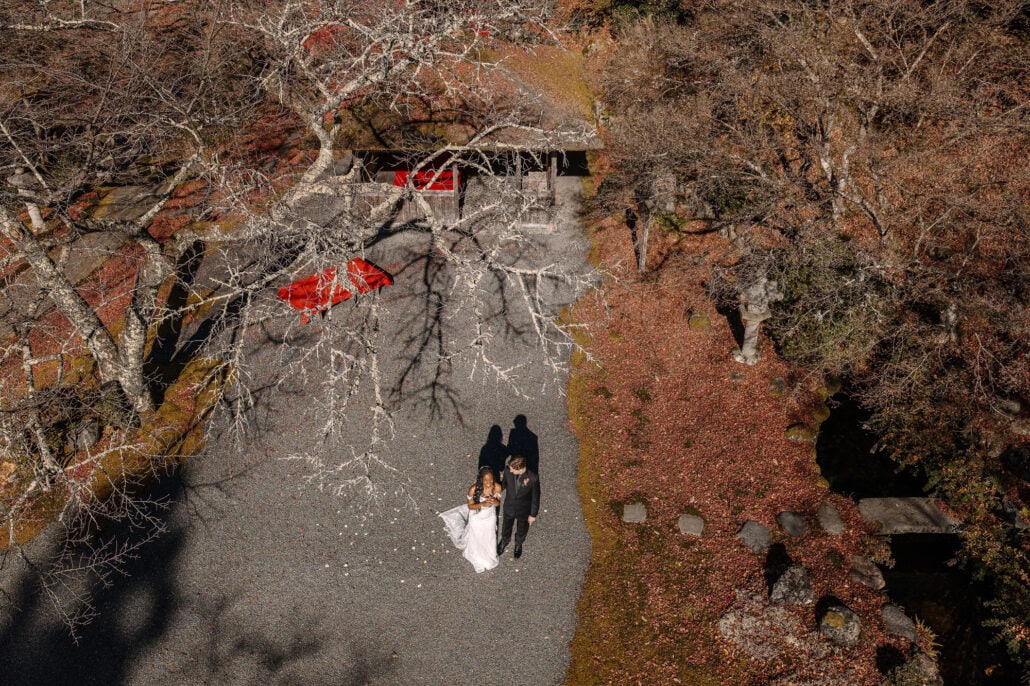 From above, the bride and groom walk a gravel path through a private garden in north Kyoto, red parasols among bare and autumn trees.