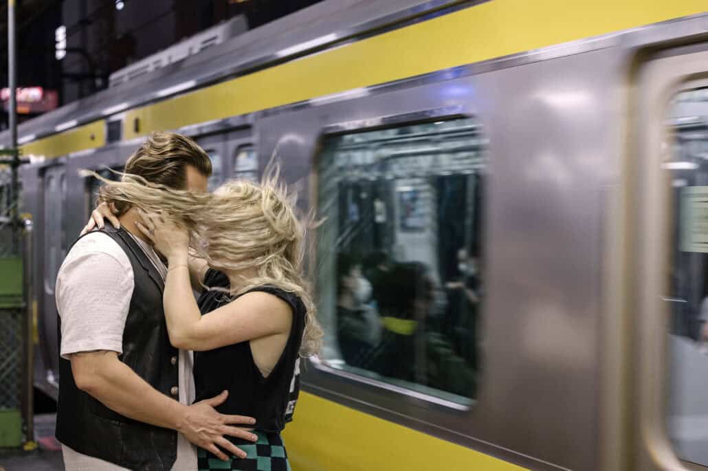 A yellow-striped Tokyo train rushes past as a couple hold each other on the platform, the city most guests arrive into when they travel to Japan for a wedding.