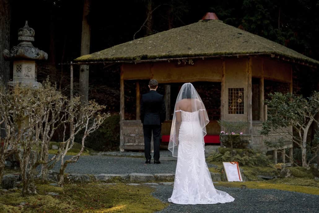 The bride walks up behind the groom, who waits facing a moss-roofed pavilion in a Japanese garden for their first look.