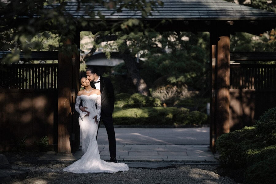 The groom kisses the bride's temple as they stand in the shade of a roofed wooden gate at a Japanese garden in Tokyo.