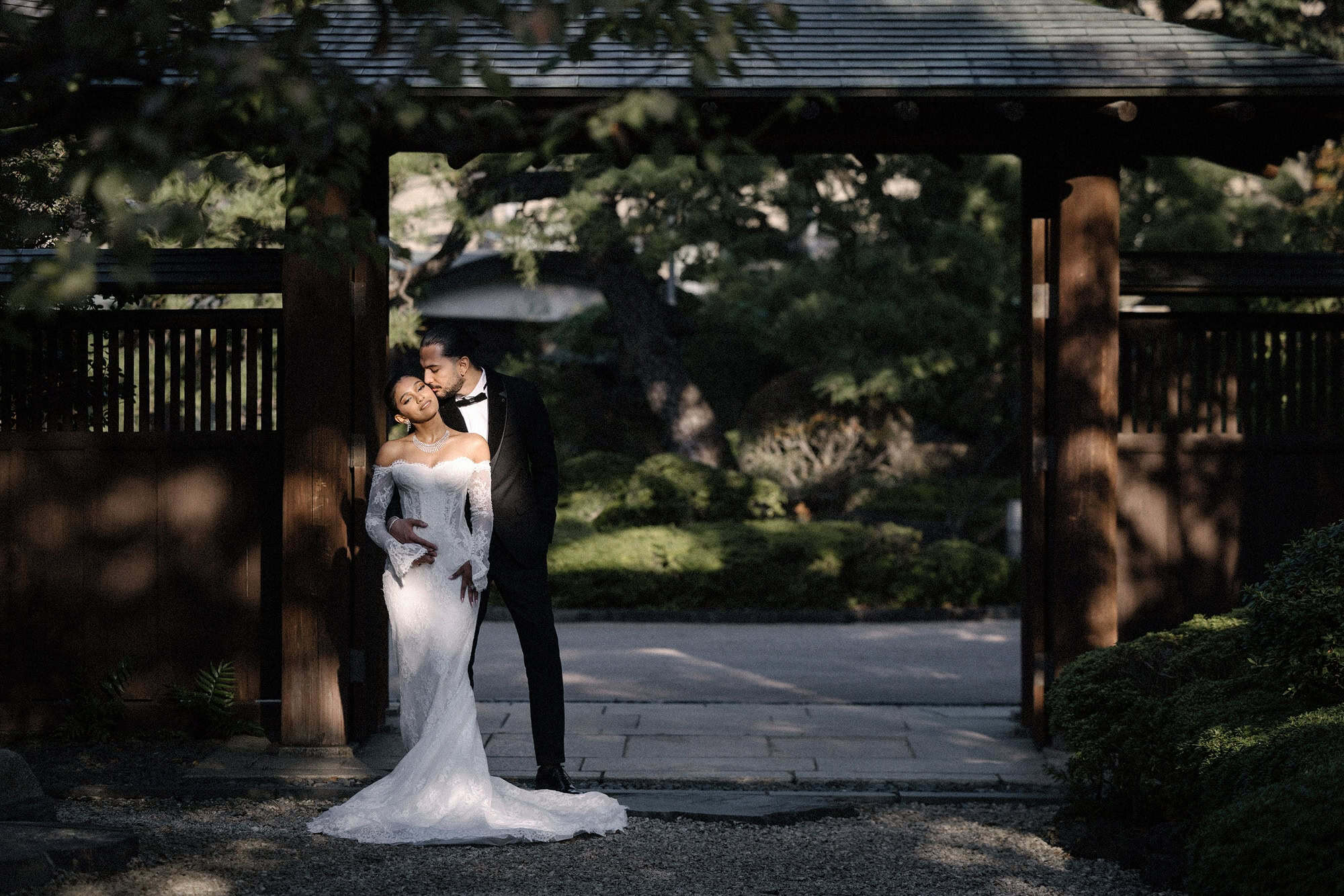 The groom kisses the bride's temple as they stand in the shade of a roofed wooden gate at a Japanese garden in Tokyo.