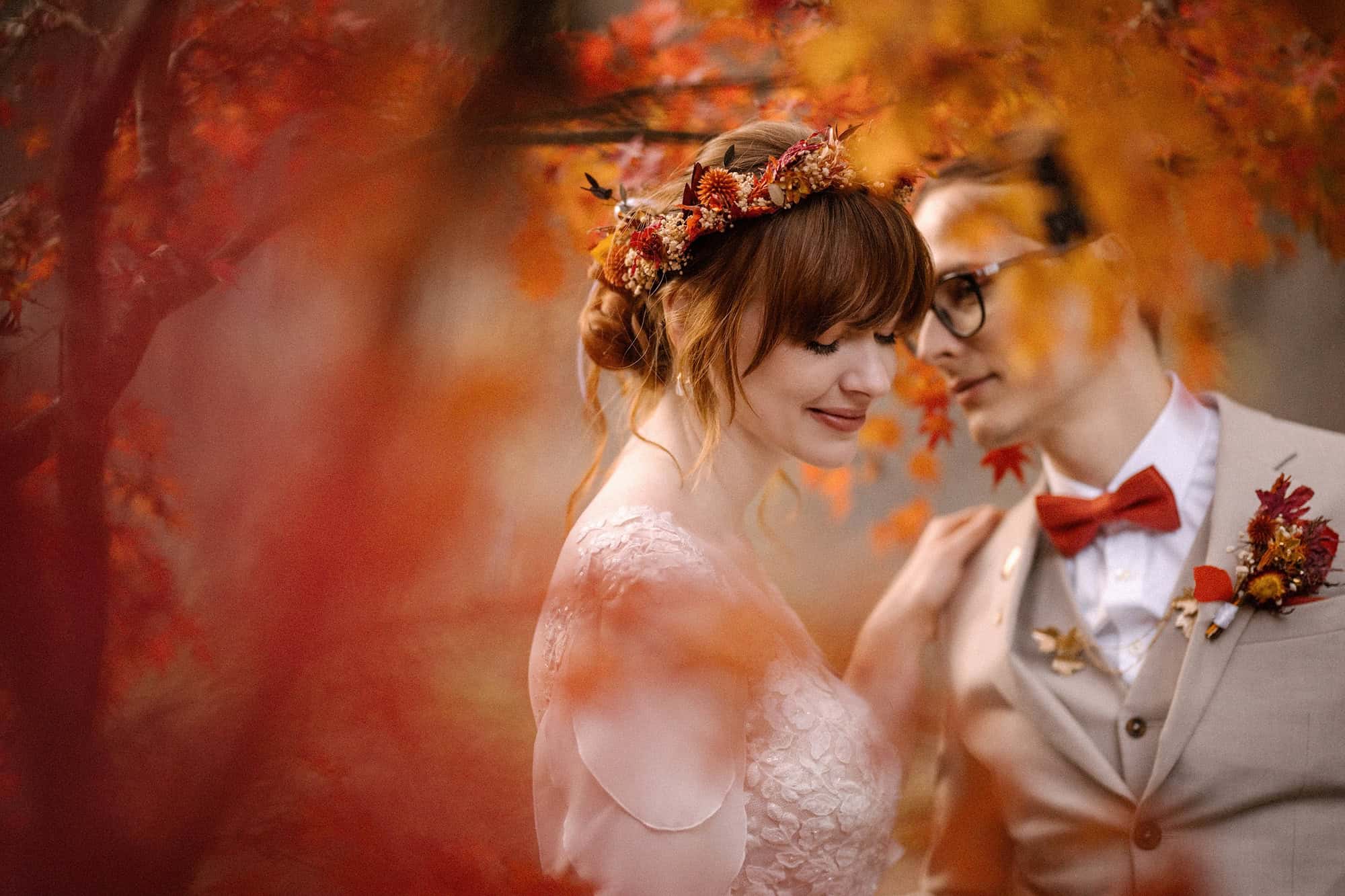 A bride and groom lean close through red and orange maple leaves at full colour, her dried flower crown matching an autumn wedding in Japan.