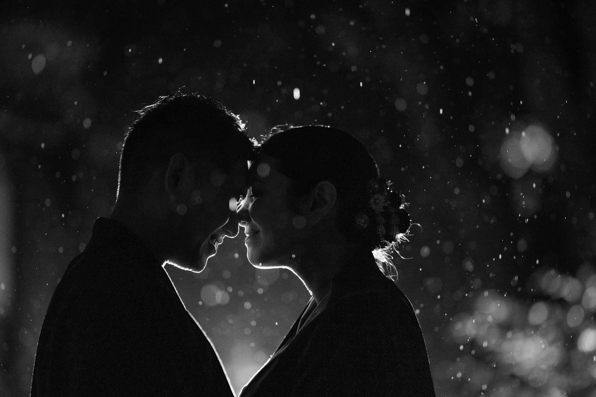 The couple stand forehead to forehead in silhouette as rain falls through the light, a night portrait in black and white.