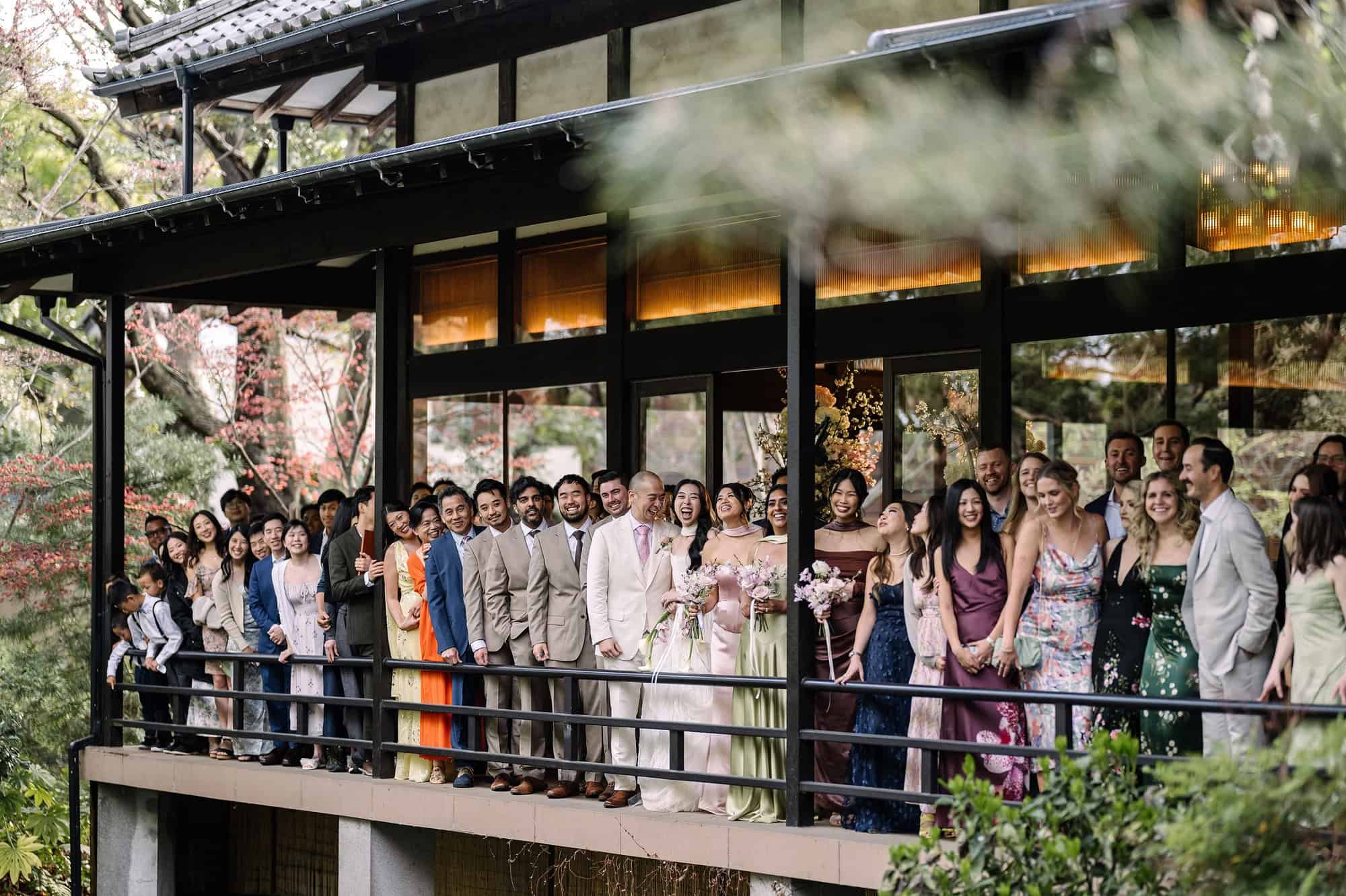 Dozens of guests line the wooden balcony of a ryokan above its garden for a group photograph, two days into a destination wedding weekend in Japan.