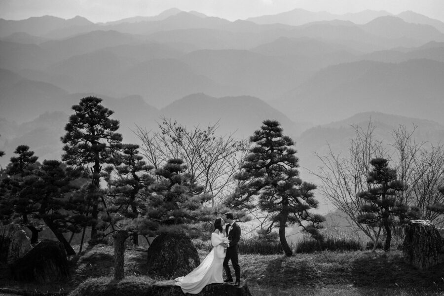 The couple stand on a flat rock among sculpted pines, layer after layer of mountains in the haze behind them, in black and white.