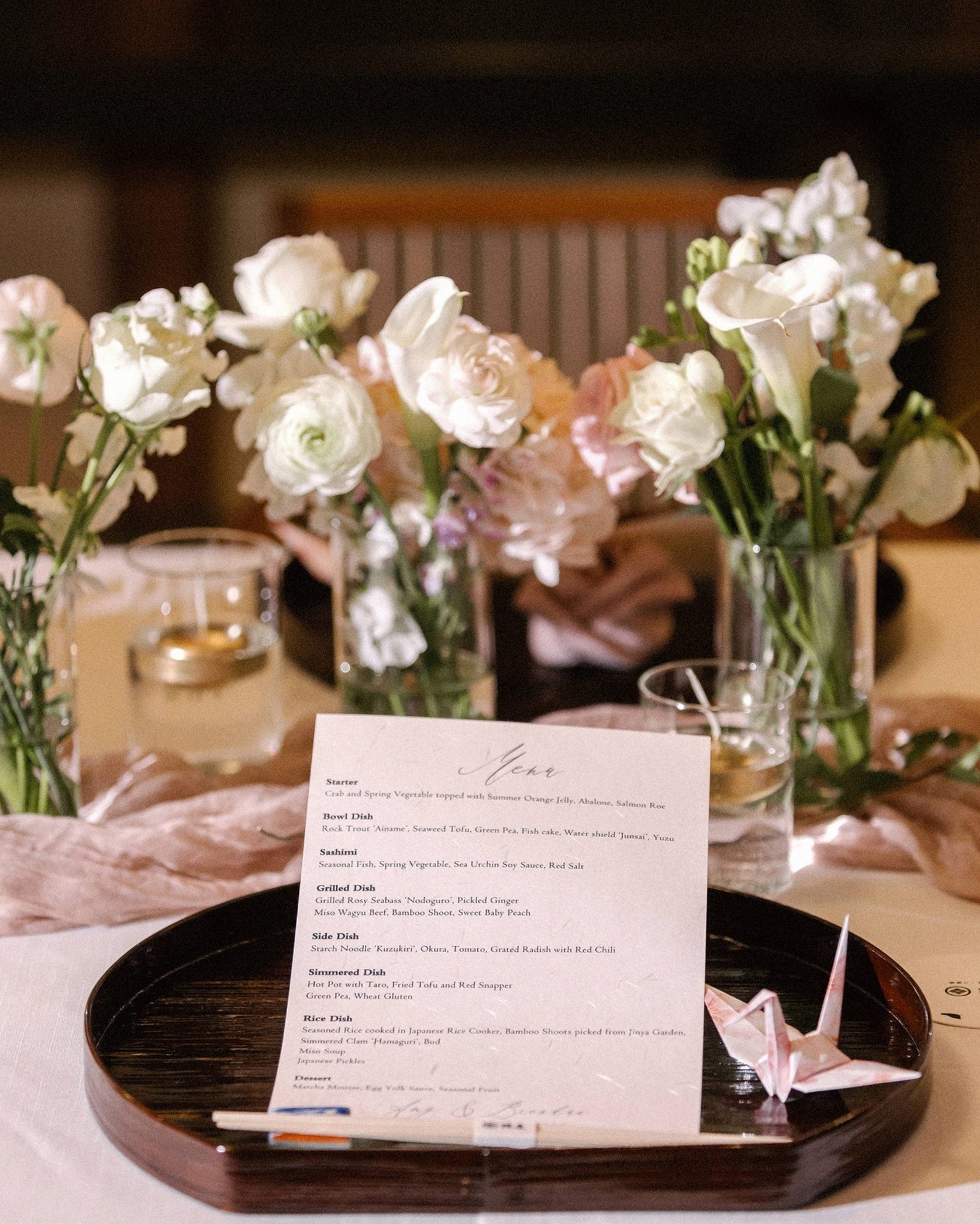 A printed wedding dinner menu on a lacquer tray with a pink paper crane, white ranunculus and calla lilies behind, at a ryokan wedding in Japan.