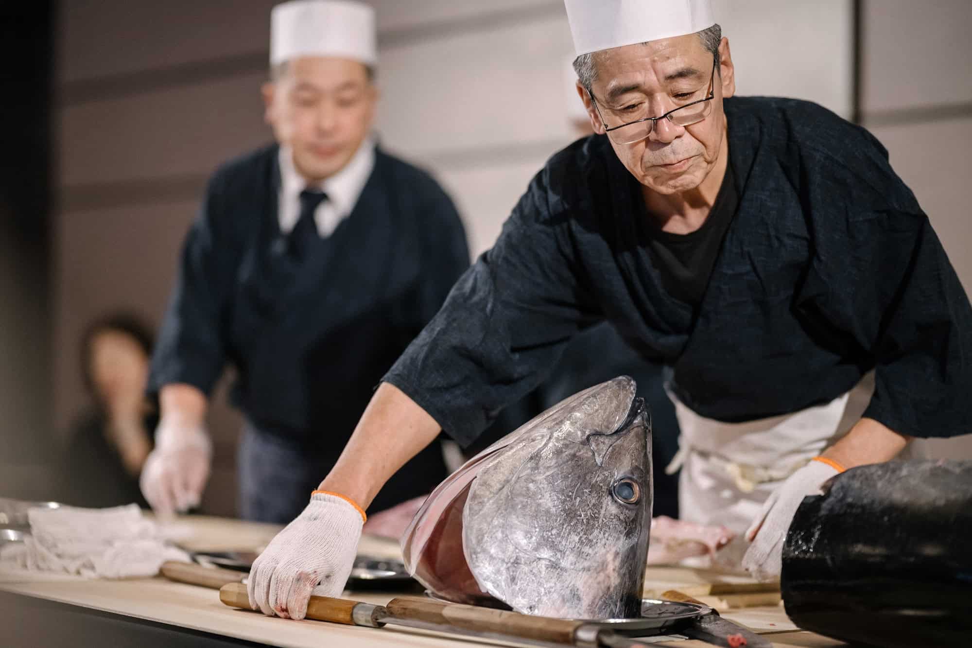 A chef in a white cap and indigo jacket breaks down a whole tuna by hand at a Tokyo wedding reception, a second chef working behind him.