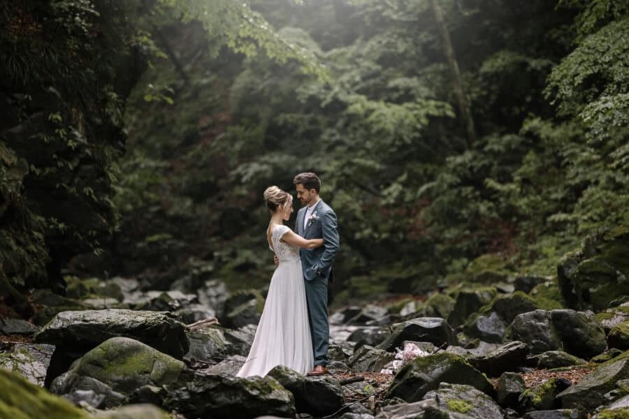 The bride and groom stand among mossy boulders in a forested mountain gorge near Kosuge, soft light falling through the trees.