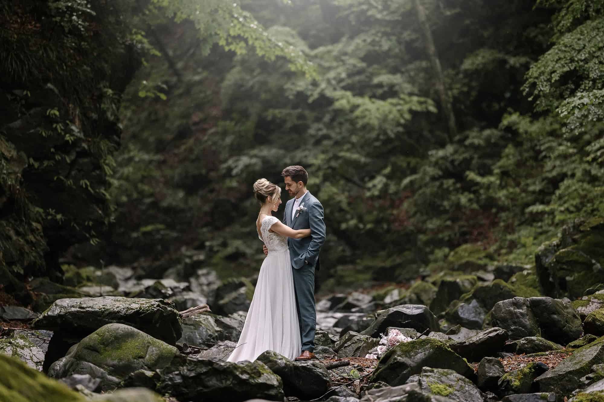 The bride and groom stand among mossy boulders in a forested mountain gorge near Kosuge, soft light falling through the trees.