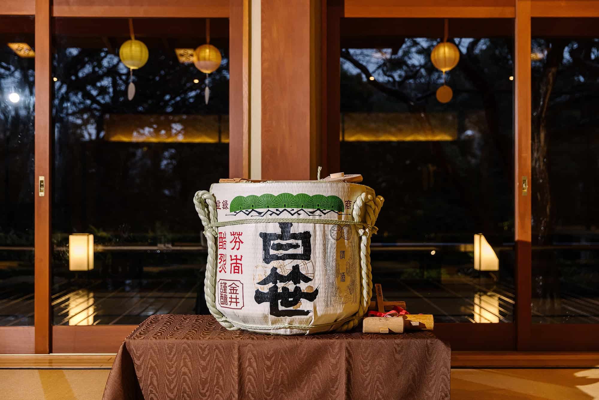 A straw-wrapped sake barrel waits on a table for kagami biraki in a ryokan hall, paper lanterns glowing beyond the glass at a welcome dinner.