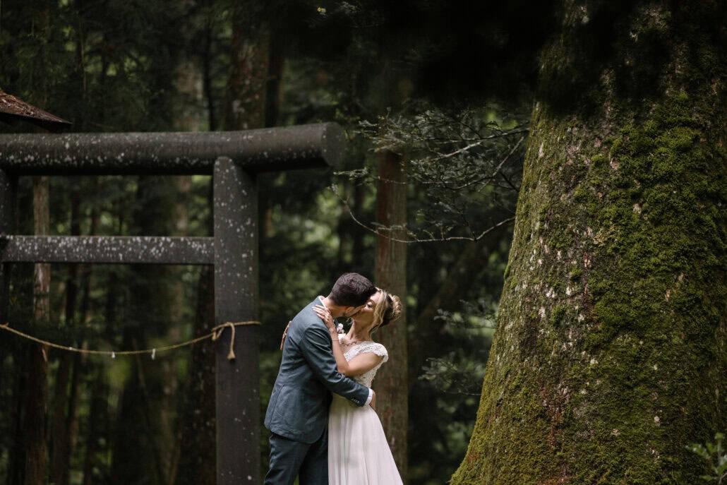 The couple kiss beside a stone torii and a huge moss-covered cedar at a small shrine in the forest, a straw rope strung between posts.