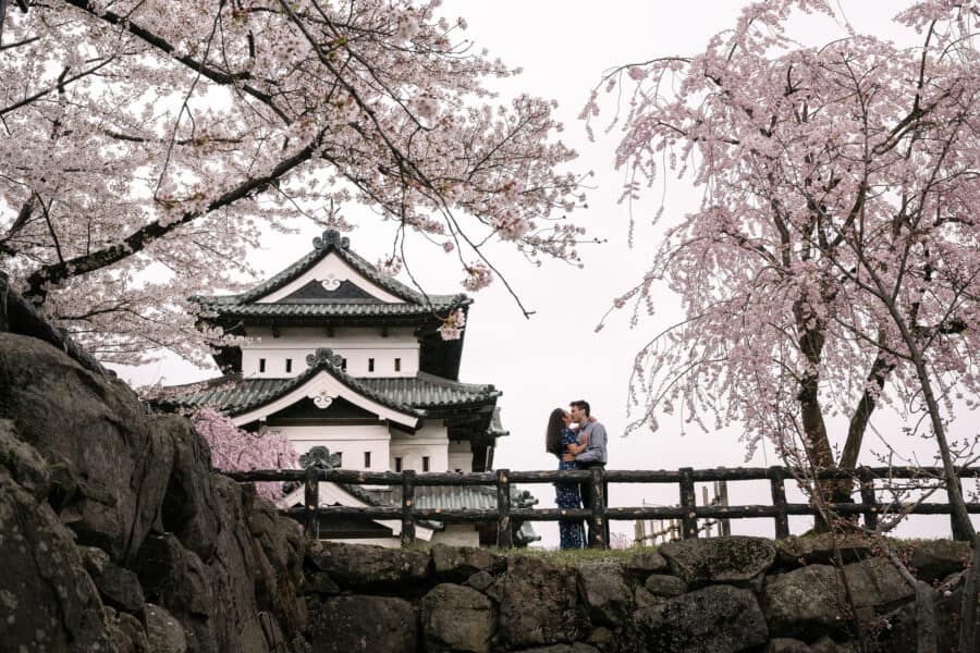 Hirosaki Castle's keep stands beyond an old stone wall as the engaged couple kiss at a wooden rail between blossoming cherry trees in Aomori.