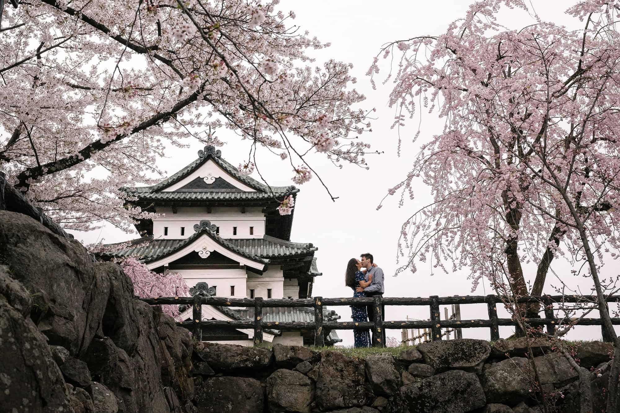 Hirosaki Castle's keep stands beyond an old stone wall as the engaged couple kiss at a wooden rail between blossoming cherry trees in Aomori.