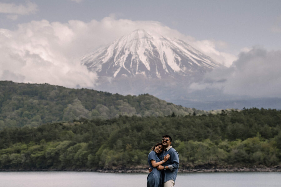 Snow covered Mt Fuji rises above forest and Lake Shoji as the couple embrace on the shore during their pre-wedding photos.