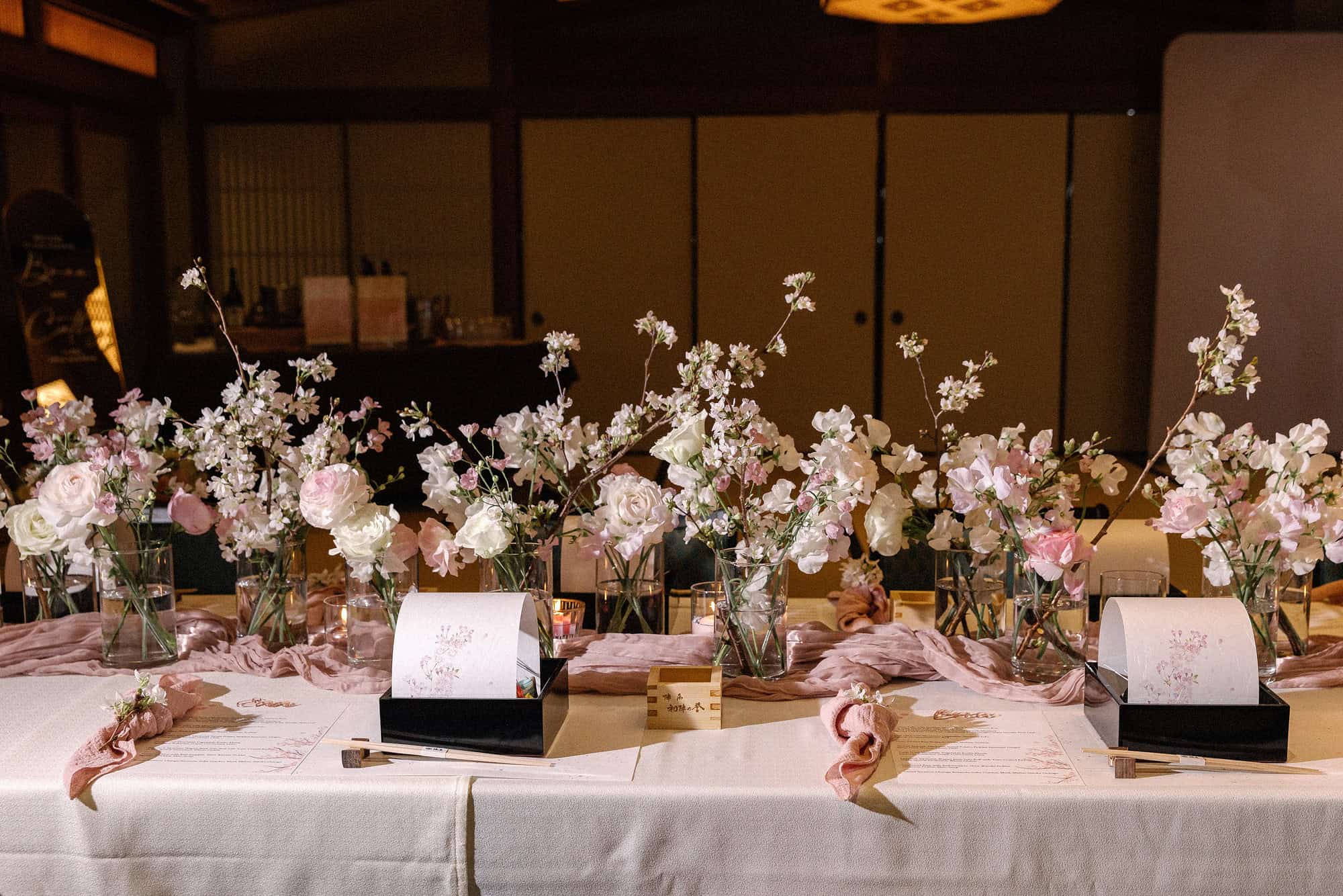 A long reception table set with cherry branches, pink roses and sweet peas, a wooden masu and blossom-printed menus, at a spring wedding in Japan.