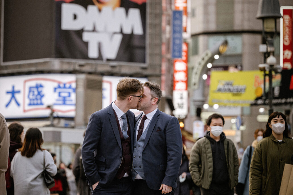 The grooms kiss on a busy Shibuya street in Tokyo at the end of their wedding day in Japan, shop signs and the crowd around them.