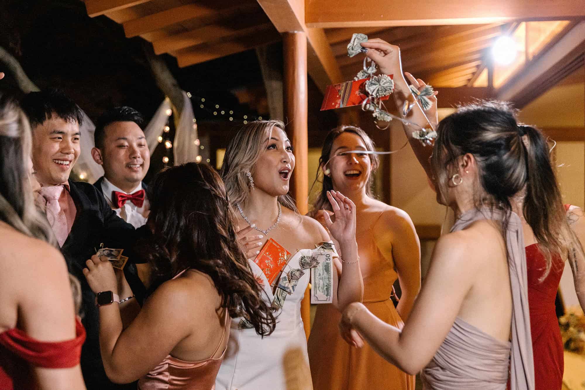 Friends laugh around a bride with folded dollar bills and a red envelope pinned to her dress during the money dance at a wedding in Japan.