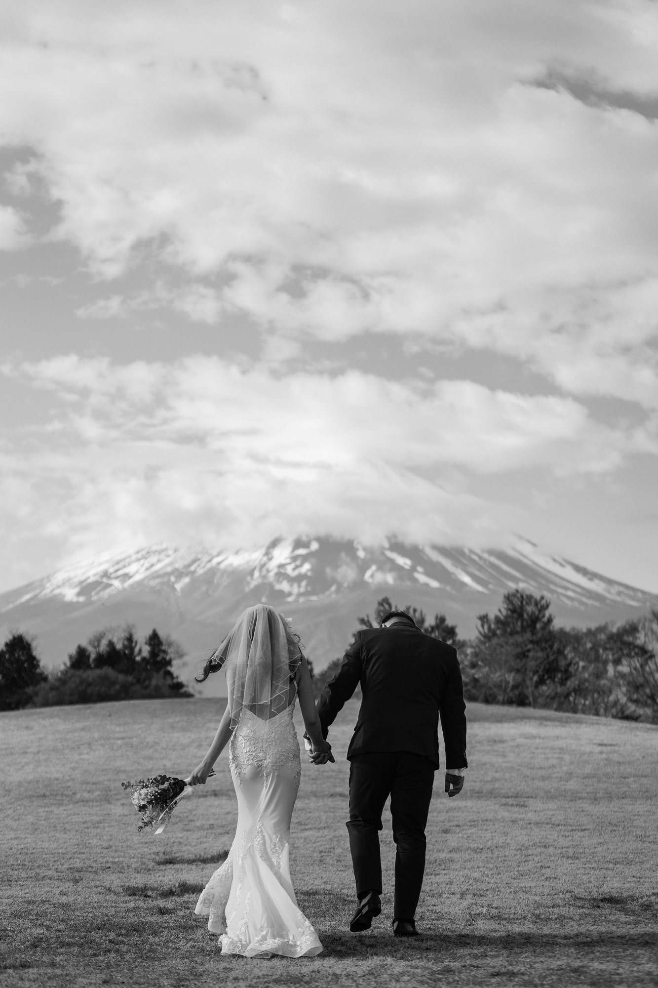 In black and white, the bride and groom walk hand in hand across an open lawn toward Mt Fuji, her veil lifting in a cool spring wind, a reminder to dress for the weather at a wedding in Japan.