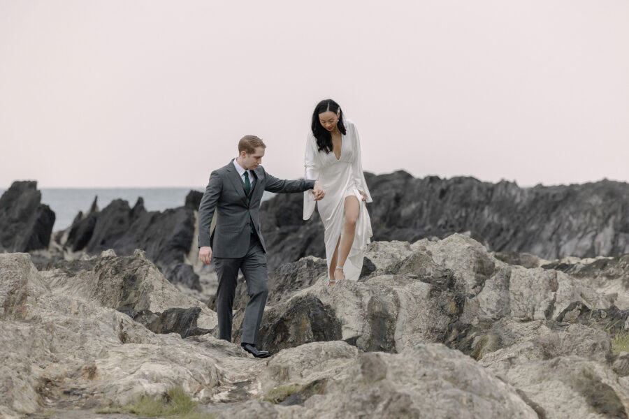 The groom helps the bride across the jagged rock layers at Arasaki Park, the sea and dark reef stretching behind them.