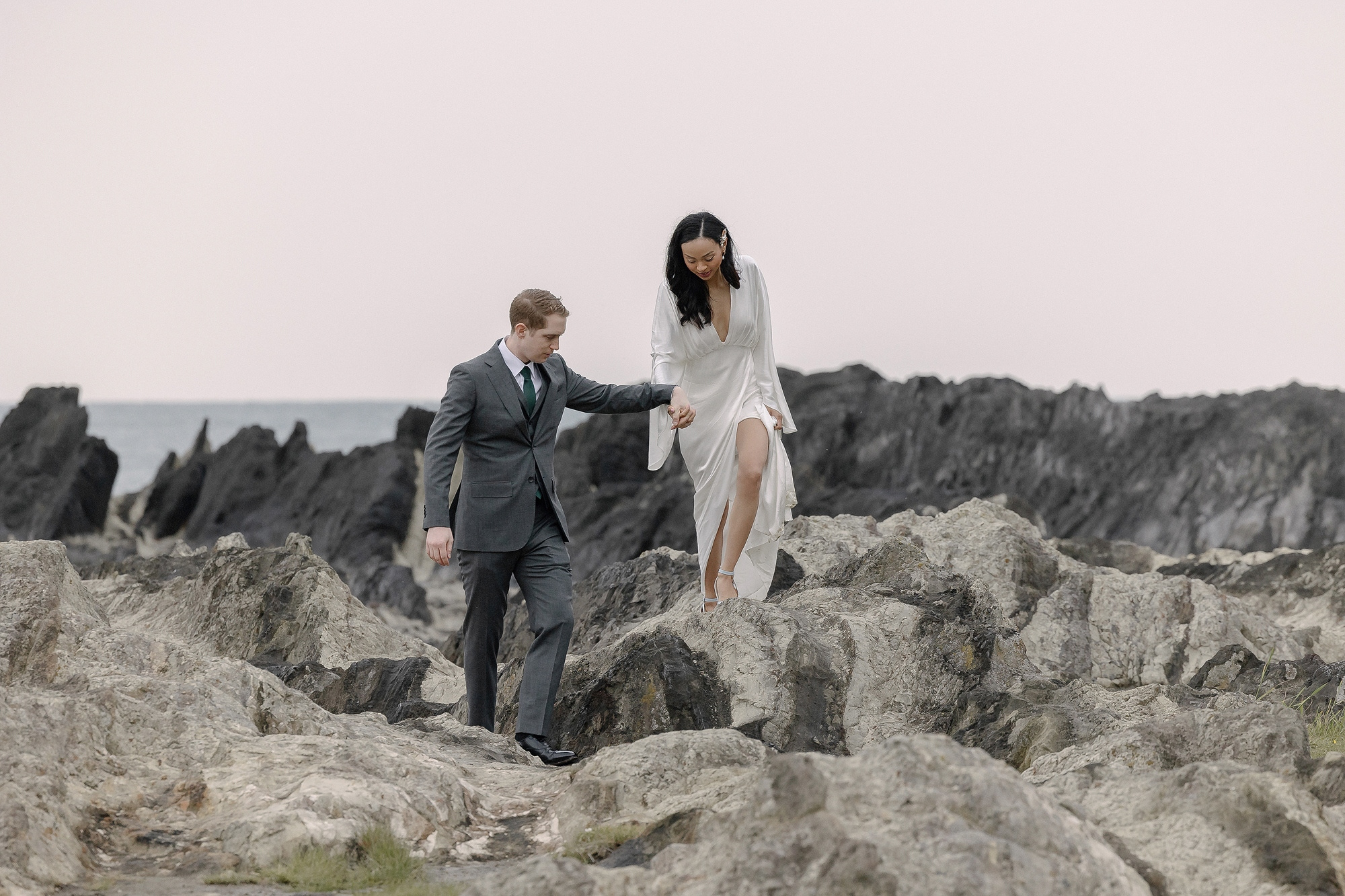 The groom helps the bride across the jagged rock layers at Arasaki Park, the sea and dark reef stretching behind them.