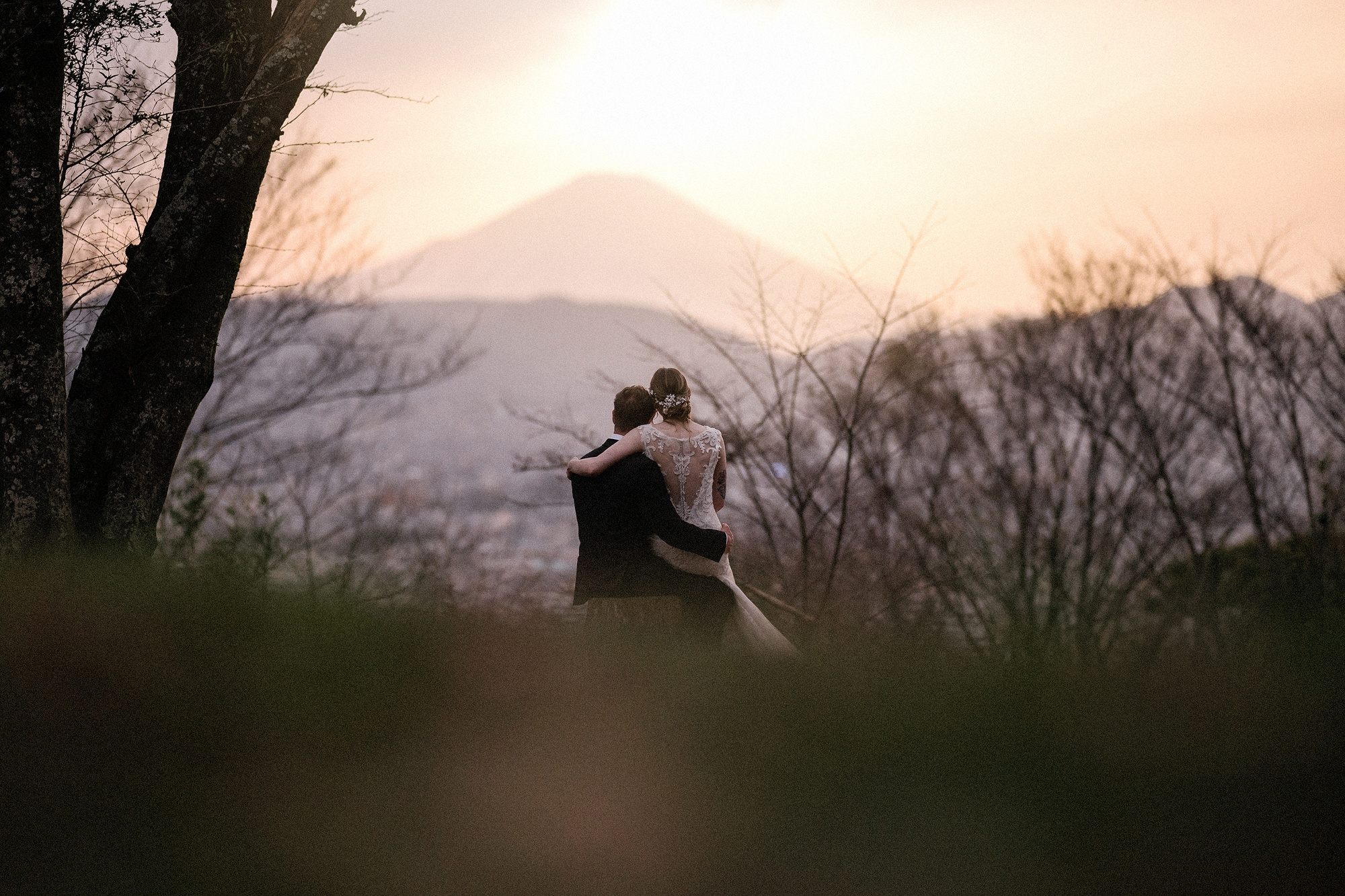 Far off on a hillside, the bride and groom embrace as the setting sun glows beside Mt Fuji, framed by trees at the close of a Japan elopement.
