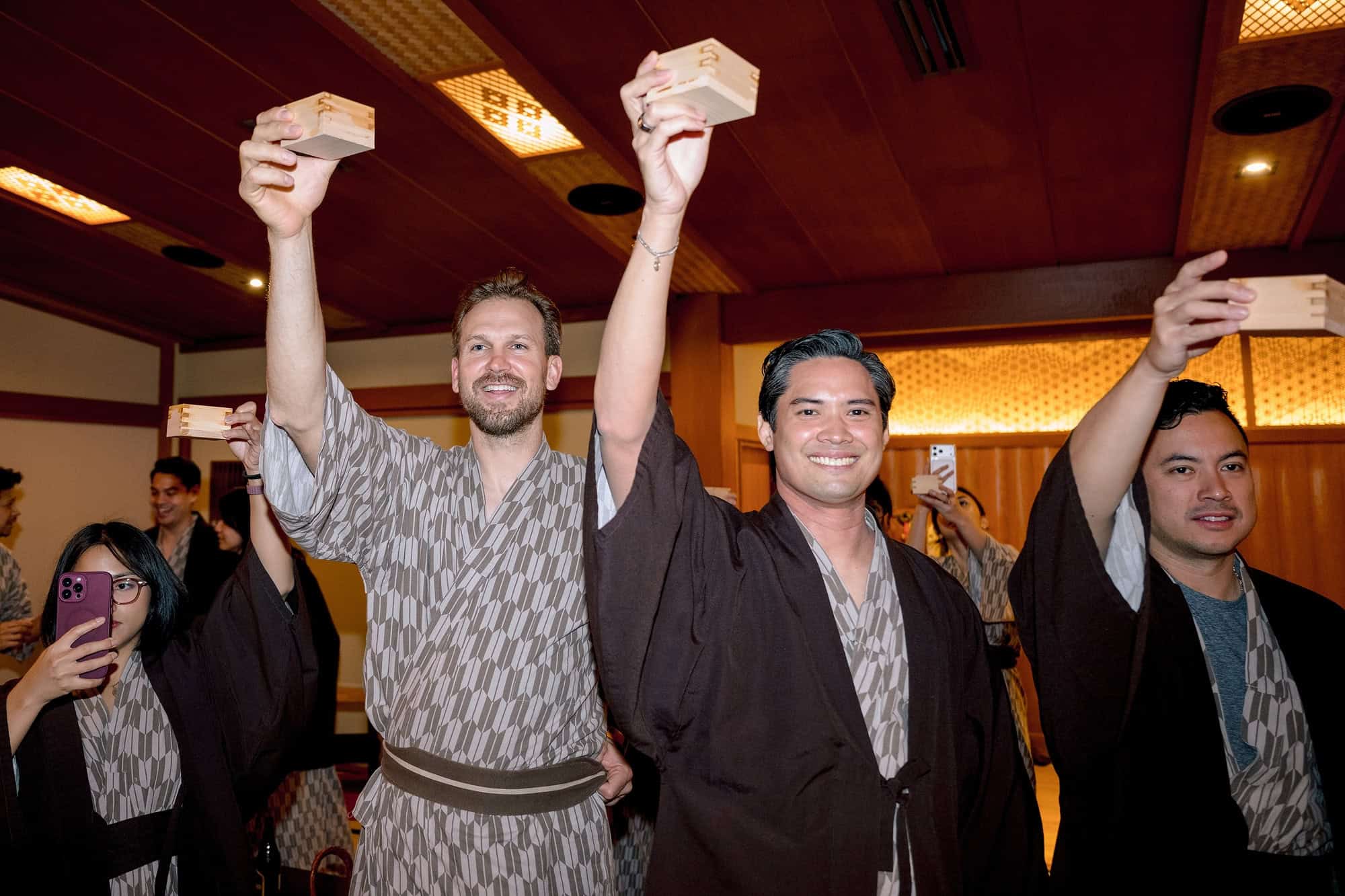 Guests in yukata raise square wooden masu cups high after kagami biraki, the sake barrel opening at a ryokan welcome dinner in Japan.